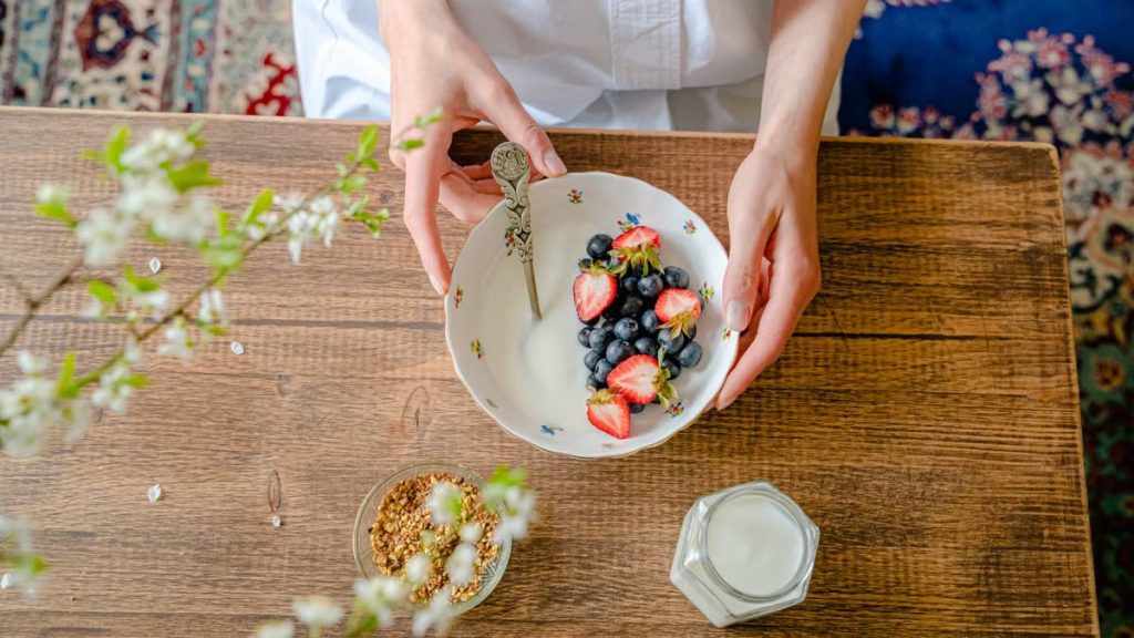 A top view of a person preparing a healthy breakfast with fresh berries, milk, and granola on a wooden table.